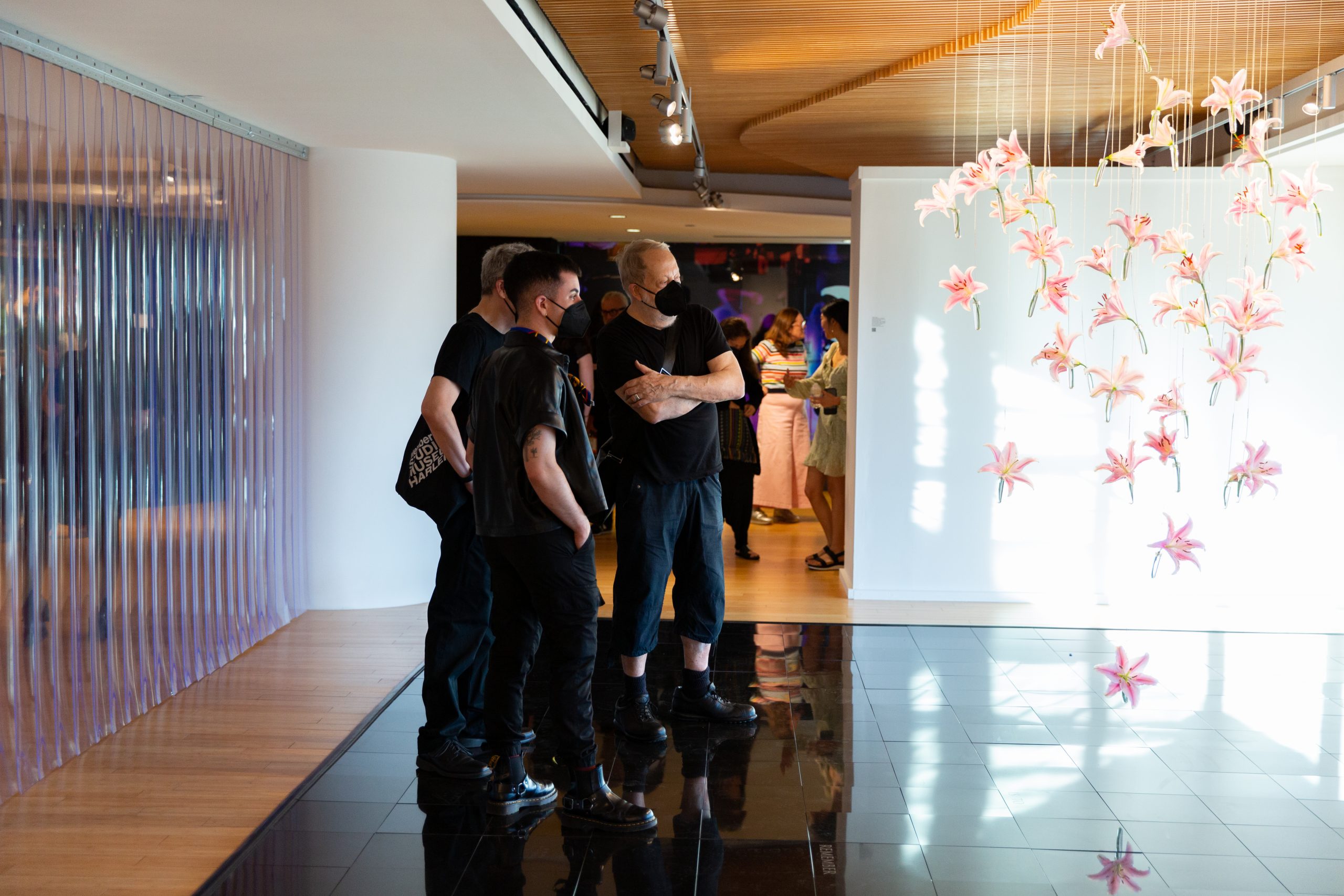 In the George Segal Gallery where the exhibition Nothing Under Heaven by Joseph Liatela is on view, three visitors stand on an installation consisting of a dance floor with black tiles and stare at the live lilies suspended from the ceiling. Behind them, to their left, is another artwork consisting of plastic strip curtains. The sun streams in the from the right and light reflects off the dance floor.