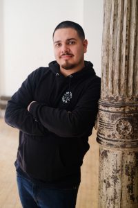 Portrait of artist Na'ye Perez wearing a black hoodie and jeans, with his arms crossed and leaning against a brown, distressed column.