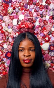 Portrait of a woman with long black hair. Behind her are bright glass flowers, ranging in varying shades of pinks and purples.