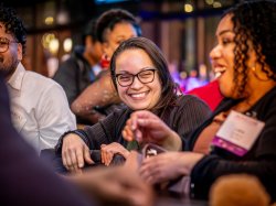 woman, seated in mid frame, wearing glasses ad smiling while a woman in foreground talks