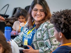"Isabella Zarate Gonzalez, wearing a blue SPARK T-shirt and a green-and-white crochet cardigan, sits at a table holding a worksheet, speaking with an elementary student who is seated around her during a classroom activity."