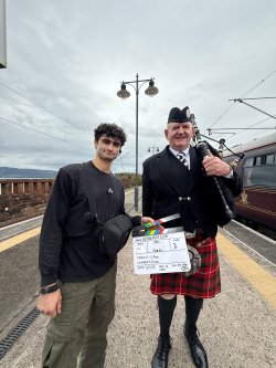 Two men standing next to each other one is holing a film set prop and another is dressed in a scottish kilt and holding a bagpipe.