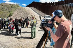 A student with headphones and a camera films a scene at the border of Mexico and Arizona.
