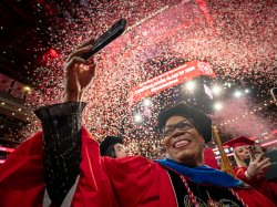 A doctoral graduate in a red gown and black doctoral cap smiles and raises a phone to capture the moment as red and white confetti rains down inside Prudential Center.