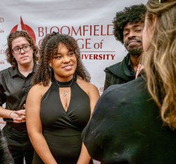 Ciara Donaldson, a 2024 Bloomfield College of Montclair State University graduate, stands at the center of the photo wearing a sleeveless black dress and a heart-shaped necklace, engaged with people gathered around her. Other individuals stand nearby, and a Bloomfield College of Montclair State University backdrop fills the background.