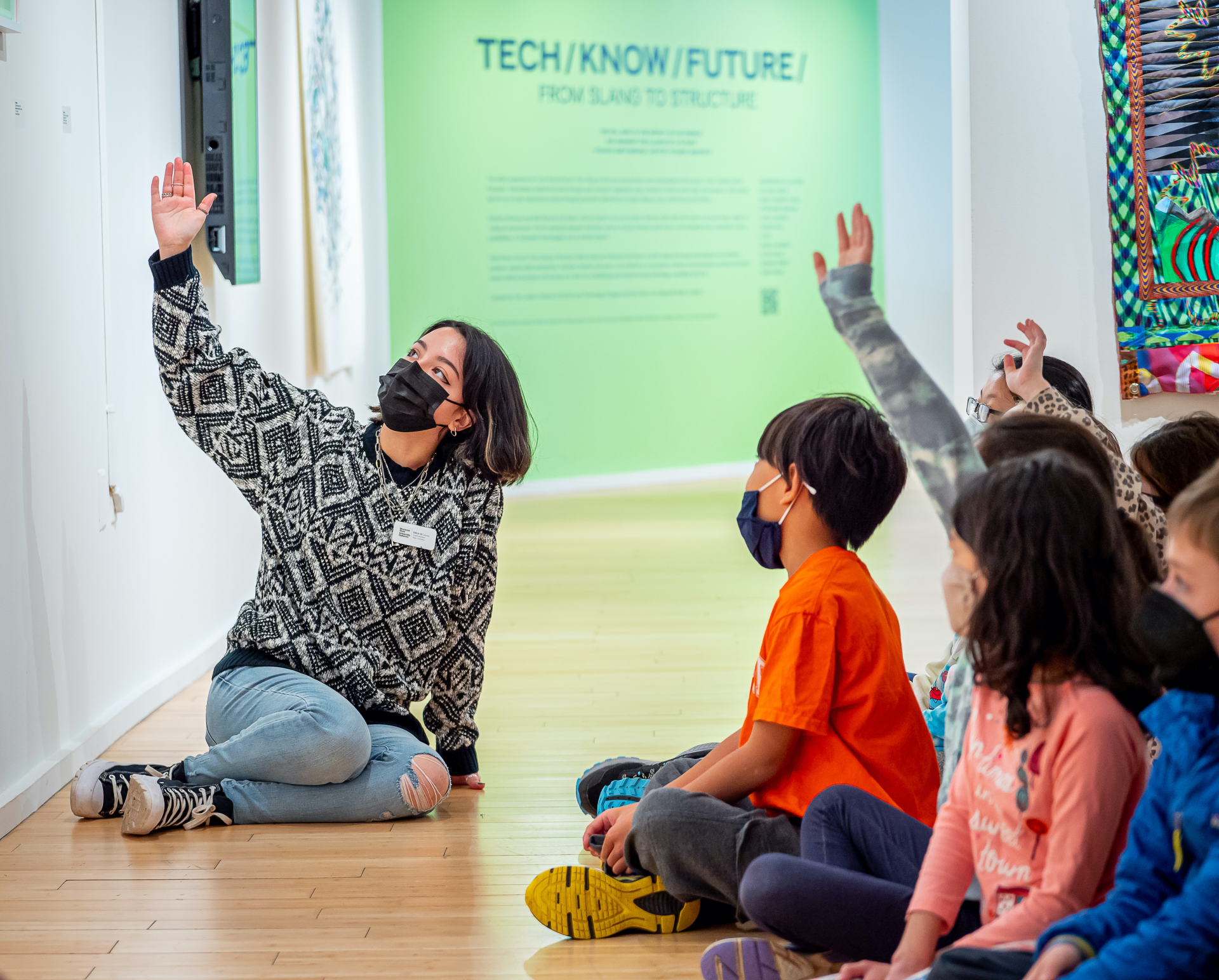 A University Galleries' student educator gestures at an artwork with their right hand. Next to them is a group of children, some of whom are raising their hands. Everyone is seated on the floor.