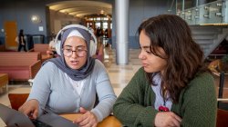 two students working together at table while one student points to a laptop