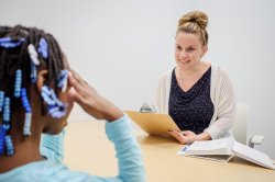 A young girl with braided hair and decorative beads sits at a table, facing an adult woman who is holding a clipboard and some papers.