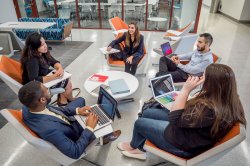 Five people are seated in a modern lounge area, around a small white table. Each person sits in an orange chair and is engaged with laptops, notebooks, or paper folders.