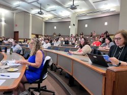 Attendees are seated at long rounded tables in a classroom.