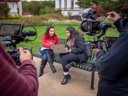 Nikki-James Soto, seated, left, films a segment of The College Tour with her friend Stephanie Grajales. A camera crew and equipment are seen in the foreground