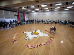 a Chakana, a revered Andean symbol, is in the middle of the floor in a large room with people sitting in a circle. A person is arranging flower petals in a circle around the Chakana