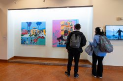 Two visitors stand and observe a colorful painting behind glass.