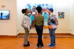 Two visitors and the artist stand before a painting behind a display case.