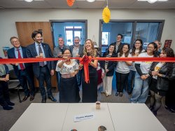 A smiling woman in a black dress cuts a ceremonial red ribbon in a classroom as spectators look on.