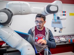 Student wearing glasses and a hoodie works on a laptop in robotics lab.