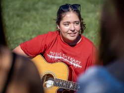 Gabriella Surace, a sophomore Music Therapy major at the John J. Cali School of Music, plays MIrrorball by Taylor Swift during a class held on the Student Center Quad. Alt Txt: A student holds a guitar while seated in the grass.