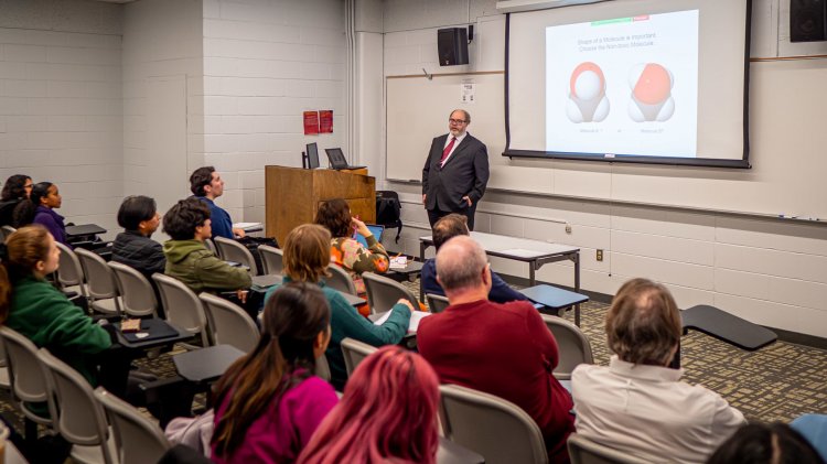 A man in a suit stands before an audience and slide presentation.