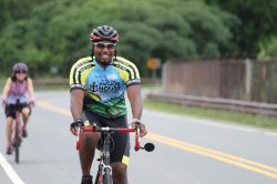 A smiling man in cycling attire that reads Anchor House rides a bicycle.