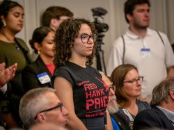 A woman in a Red Haw Pride t-shirt stands in the back of the hearing room.