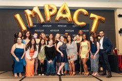 A group of students and staff stand onstage under the word IMPACT.