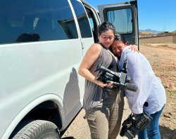 A student holding a video camera hugs another student in the desert.