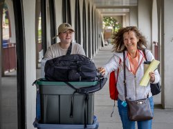 Alt Text: A parent and student push a cart loaded with college essentials into a dorm room.