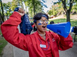 A student wearing a red Montclair State University jacket holds a bullhorn and smiles while directing new students during Welcome Week festivities.