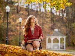 Student holds a pencil and pad while sitting on Montclair's campus. The fall foliage is orange and. yellow.