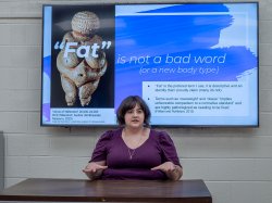 Stepahnie Spitz gestures while seated at a desk with a slide of Venus of Willendorf and “fat is not a bad word” projected behind her.