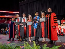 The Povolo quintuplets, wearing graduation caps and gowns, stand together on stage at Montclair State University's 2024 Commencement ceremony. Each holds their diploma while smiling alongside university officials in academic regalia.