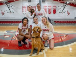 Members of the Montclair State University women's basketball team pose together on the basketball court with two dogs: Pebbles, a small brown labradoodle, being held by one of the players, and Charley, a golden retriever, sitting in front and looking at the camera.