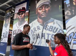 A young reporter interviews a New Jersey Jackals baseball player in front of a display featuring historical baseball players at the Hinchliffe Stadium exhibit.