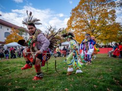 Native American men and women in traditional costumes dance outdoors while spectators watch.