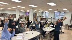 adult students seated at four tables in a library, stretching arms upward following the teacher at the front of the room