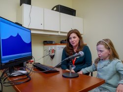 8-year-old Colette Fougere says ‘r’ into a microphone while Alexis Gallagher looks on.