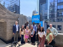 A group of students and communications professionals standing on a New York City rooftop in two rows. Everyone is smiling and happy.