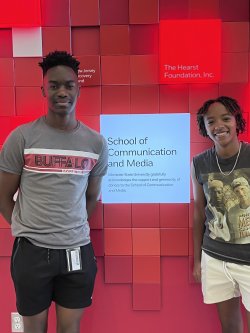 Two students smiling and standing in front of a red background in a college building.