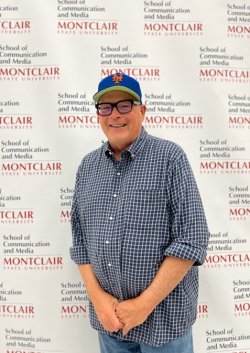 A man standing in front of a backdrop with writing. He has glasses, a blue hat and is wearing a button down dress shirt with jeans.