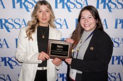 Two college students dressed professionally standing in front of a white backdrop with blue lettering. They are holding an award.