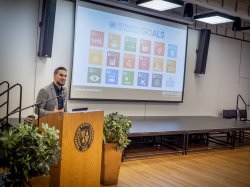 Director of Sustainability, Benjamin Kalscheur, standing at a podium giving a presentation. The projector screen behind him shows a graphic of the United Nation Sustainable Development Goals.