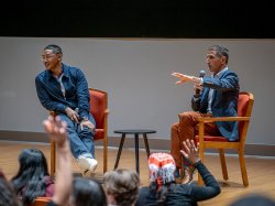 A man gestures as two men sit onstage during a Q&A with students.