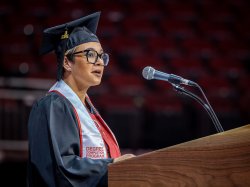 Female degree completion student speaking at Commencement in black regalia with white "Degree Completion Program" graduation stole.