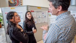 Two young scientists talk to a professor in front of a research poster.