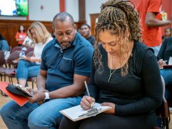 Two parents sitting next to each other, writing a letter on a clipboard.