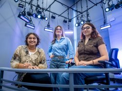 In a newsroom, three women, two seated at a desk and one standing between them pose with studio lights shining behind them.