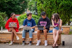 Four students sitting on a low brick wall on a sunny green quad. One girl is holding up her phone and gesturing toward it while the other three, using their laptops, look over at what she's showing them.