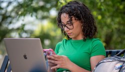 Woman on bench outdoors on a sunny day checks her phone while using laptop