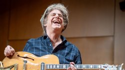 Pat Metheny laughs on stage as he holds a guitar in his lap.