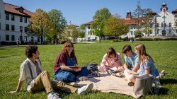 Students sit together on a blanket on a large grassy lawn at Montclair State University, talking and studying with notebooks and a laptop open, while academic buildings with red-tiled roofs and a clear blue sky frame the background.